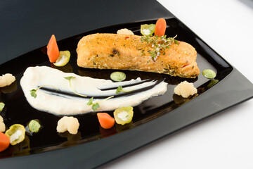 Fillet of red fish with vegetables, herbs and white sauce on a black glossy plate. Gourmet restaurant dish.