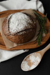 Round black bread on baking paper and a wooden spoon with flour on a black background.