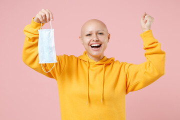 Young bald happy woman 20s without hair in yellow casual shirt hold sterile face mask, safe coronavirus covid-19, quarantine do winner gesture clench fist isolated on pastel pink background studio.