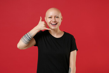 Young bald smiling caucasian positive tattooed woman 20s without hair wearing black basic casual t-shirt doing phone gesture like says call me back isolated on red color background studio portrait.