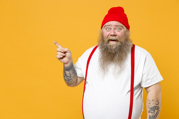Fototapeta na wymiar Fat pudge obese chubby overweight tattooed bearded surprised man shocked has big belly in white t-shirt red hat suspenders point index finger aside on workspace isolated on yellow background studio.