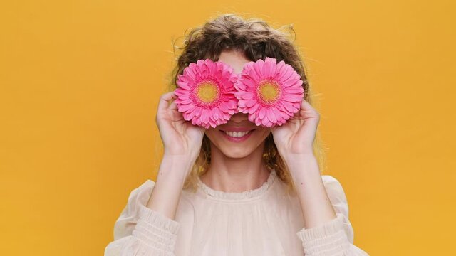 Dancing Happy Young Woman Smiling Twirls Pink Flowers Near her Eyes Opens Face Sticks out Tongue on Yellow Background. Portrait of Glamorous, Beautiful Sweet Girl Positive Emotions people. Fashion