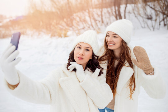 Concept friendship lifestyle. Two beautiful young woman with white hats taking selfie photo with smartphone outdoors in winter
