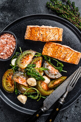 Fish meal with Roasted Salmon Fillet Steaks and arugula tomato salad on a plate. Black background. Top view