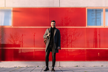 Young businessman with backpack smiling in front of a red wall at sunset looking at the camera.