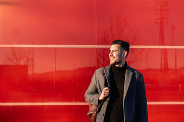 Young businessman with backpack smiling in front of a red wall at sunset