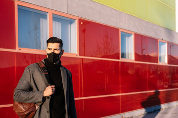 Young businessman with backpack and face mask in front of a modern building at sunset