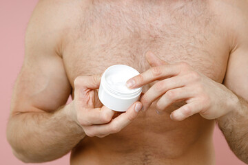 Cropped image of naked young man 20s perfect skin applying moisturizer facial cream from container isolated on pastel pink background studio portrait. Skin care healthcare cosmetic procedures concept.