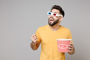 Young man in 3d glasses yellow t-shirt watch movie film, hold bucket of popcorn clench fist winner gesture look aside isolated on grey background. People sincere emotions in cinema, lifestyle concept.