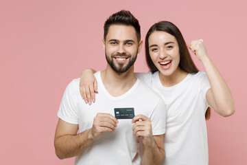 Young lovely couple two friends bearded man brunette woman in white basic blank print design t-shirts hold in hands mockup of credit bank card isolated on pastel pink color background studio portrait.