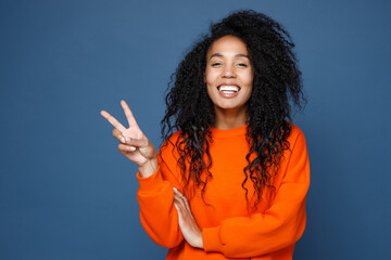 Cheerful laughing funny young african american woman wearing casual basic bright orange sweatshirt standing showing victory sign looking camera isolated on blue color wall background studio portrait.