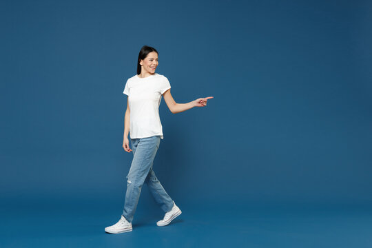 Full length young smiling latin woman 20s wearing white casual basic t-shirt point index finger aside back on copy space mock up area workspace isolated on dark blue color background studio portrait.