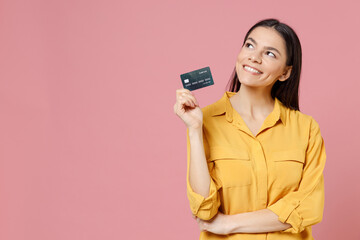 Naklejka premium Young latin happy dreamful pensive wistful cute attractive satisfied rich successful woman 20s in yellow shirt hold credit bank card look aside isolated on pastel pink background studio portrait.