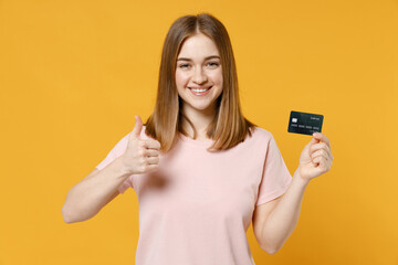 Young smiling happy rich student woman 20s in casual basic pastel pink t-shirt blank print design holding credit bank card show thumb up like gesture isolated on yellow background studio portrait.