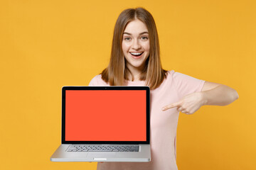 Naklejka premium Young surprised freelancer woman 20s wearing casual basic pastel pink t-shirt point index finger on laptop pc computer with blank screen workspace area isolated on yellow background studio portrait