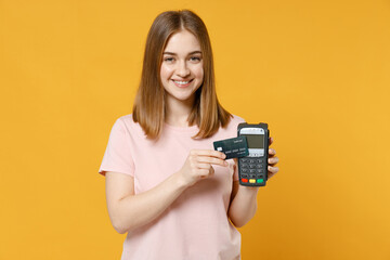 Young woman in basic pastel pink t-shirt hold wireless modern bank payment terminal to process and acquire bank credit card payments show ok okay gesture isolated on yellow background studio portrait.