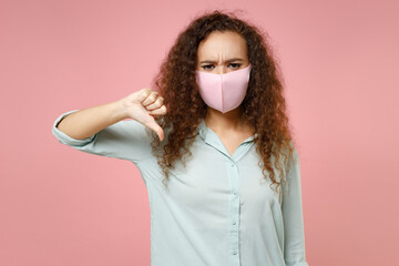 Young black african curly woman in blue shirt reusable face mask safe from coronavirus virus covid-19, pandemic quarantine show thumb down gesture isolated on pastel pink background studio portrait