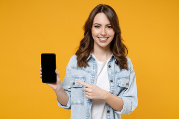 Young smiling happy fun woman 20s wear stylish casual denim shirt white t-shirt point index finger on mobile cell phone with blank screen workspace area isolated on yellow background studio portrait