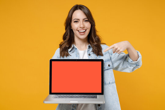 Young smiling happy satisfied fun woman 20s in denim shirt white t-shirt point index finger on laptop pc computer with blank screen workspace area chat isolated on yellow background studio portrait