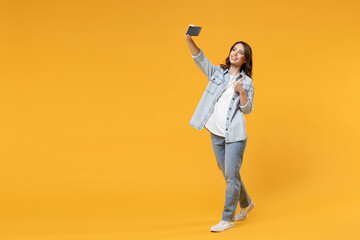 Full length of young smiling friendly fun student woman in casual denim shirt white t-shirt doing selfie shot on mobile phone show thumb up like gesture isolated on yellow background studio portrait.