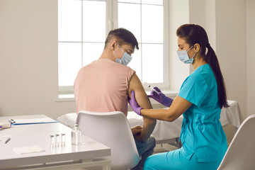 Female medical worker in blue uniform sitting making injection of coronavirus vaccination for caucasian man patient in face mask during pandemic in medical clinic. Vaccine against COVID-19 infection