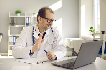 Happy physician sitting at office desk and waving hello at laptop computer greeting patient in video conference meeting. Online doctor, telemedicine consultation, virtual visit to hospital concept