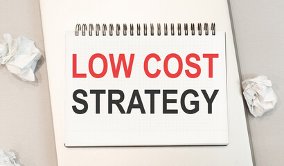 The word low cost strategy is written in a notebook that sits on a gray desktop along with a laptop.