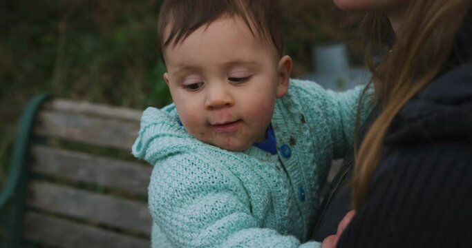 A little baby is sitting on a bench outdoors in the garden with his mother