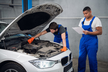 A mechanic in blue overalls examines a broken engine of a car. His partner takes notes