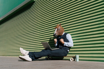 Businessman in horse mask with laptop gesturing while sitting against green wall