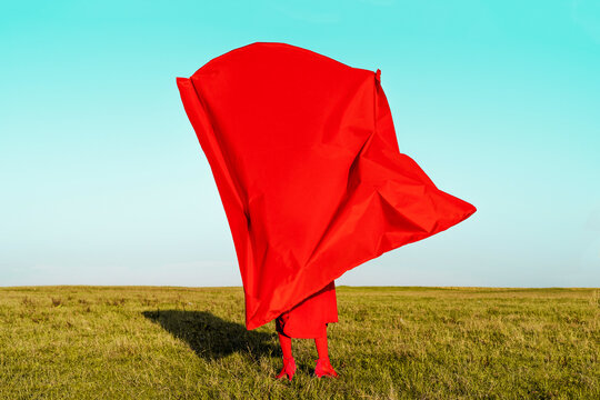 Woman covering herself with red fabric while standing on field against blue sky