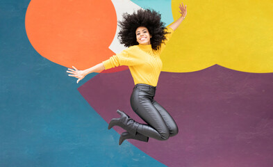 Cheerful woman jumping with arms outstretched against colorful wall