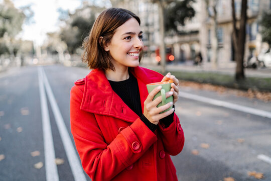 Smiling young woman holding coffee cup while looking away on street