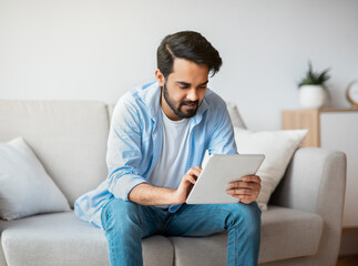 Fototapeta premium Handsome Eastern Guy Using Digital Tablet While Relaxing On Couch At Home