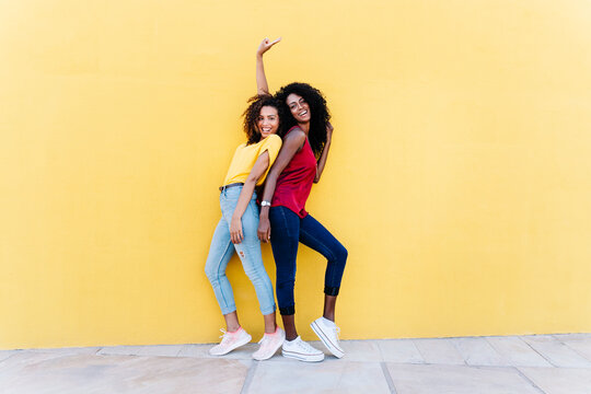 Cheerful girlfriends enjoying while standing back to back by yellow wall