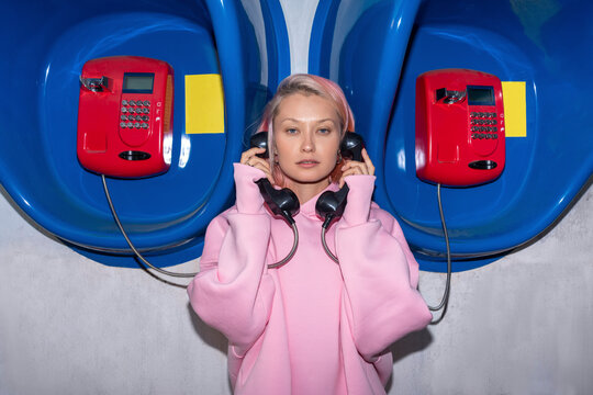 Young woman with pink hair wearing pink hooded shirt standing in front of telephone booths, holding receivers