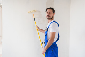 A bearded builder in work clothes paints the wall with a roller. He smiles friendly and poses for the camera