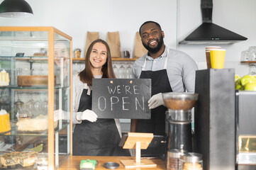 Cheerful caucasian female barista and african american male barista standing behind a countertop in cozy cafe, hold open sign board together. Small business opening