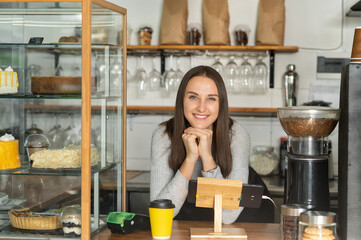 Friendly and smiling waitress in apron leaned on the counter bar and looks at the camera and smiles. Cheerful female bakery owner