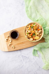Top View of Bowl of Tofu Cashew Fried Rice with a Small Bowl of Soy Sauce and Cashews on a Board; Green Material in Background; White and Gray Marble Countertop