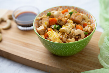  Bowl of Tofu Cashew Fried Rice with a Small Bowl of Soy Sauce and Cashews on a Board; Green Material in Background; White and Gray Marble Countertop