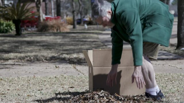 A senior adult homeowner working in his yard picking up a pile of raked leaves on a cool sunny morning.