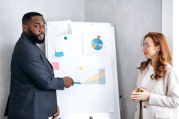 African American man in stylish suit presentation work report on conference with smiling caucasian lady. Influential business partners standing near flipchart with financial graphs, talking about