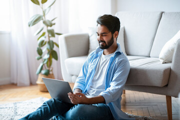 Telecommuting Concept. Young Eastern Guy Freelancer Working On Laptop In Living Room