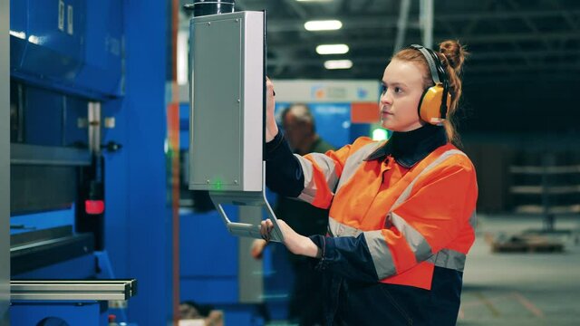 Female worker in ear protectors uses portable screen to operate a machine