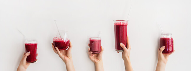 Human hands holding glasses with purple beet pomegranate fresh homemade smoothie with glass straws, white background, wide composition. Detox, dieting, weight loss, healthy lifestyle concept - 417960744