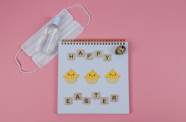 Notepad and mask.
Notepad with chickens and the words happy easter made from wooden cubes with a mask and disinfectant on a pink background, top view close-up.