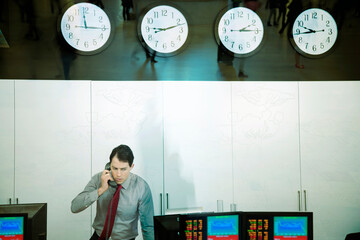 Male stock trader talking on the phone in a trading room with multiple clocks and computer screens