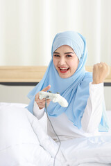 VDO game console station concept. active asian muslim woman wearing white sleepwear sitting on bed, holding joystick and playing exciting game. Cute girl looked excited with game controller console