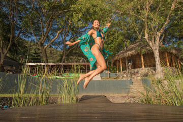 Latin woman jumping on a pier, she is smiling, she is on vacation, enjoying, a tropical landscape with trees and a palm hut behind, on a sunny summer day